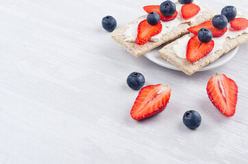 Summer light sweet vegetarian open sandwiches on crisps bread with ripe strawberry slices, blueberry, cream cheese on soft light white wood board, closeup, copy space.