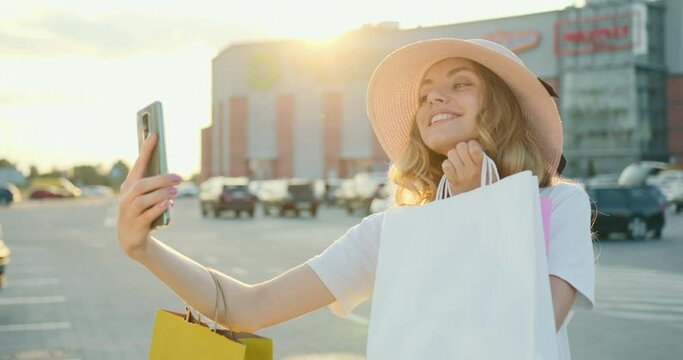 A girl is taking a selfie on her smartphone. She is smiling and holding the shopping bags in her hands. Shopping center in the background. 4K