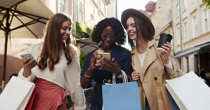 Happy multi-ethnic girls walking outdoors after shopping. Beautiful African American woman with tapping on smartphone and showing it to girlfriends. Caucasian female with cup of coffee. Fun concept