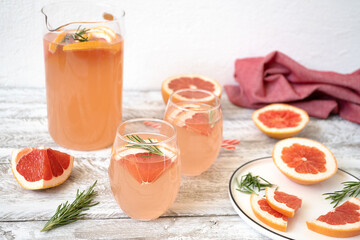 Summer cocktail with grapefruit slices and sprigs of rosemary in a jug and glasses stands on a vintage wooden table. Nearby is a white bowl with grapefruit slices and fresh rosemary. 