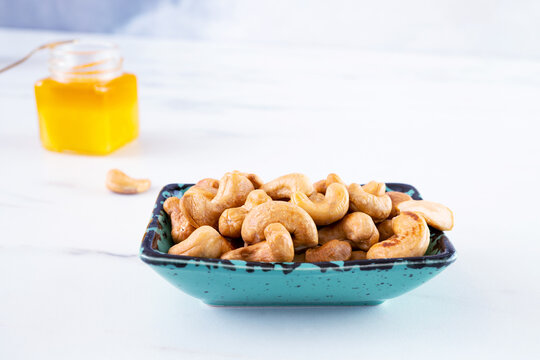 A Rectangular Blue Bowl Of Whole Roasted Cashews Stands On A White Table. A Glass Jar With Honey Stands In The Background. Delicious And Healthy Snack, Natural Organic Products. Selective Focus.