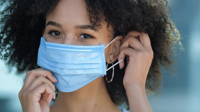 Beautiful smiling young african american woman putting mask on face to protect from flu coronavirus outbreak second wave of pandemic, looking at camera outdoor. Prevention of disease in public places