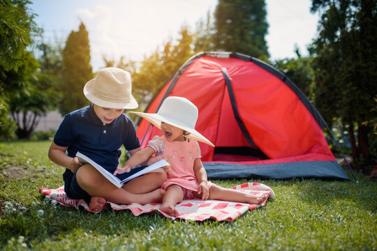 Two happy little kids, boy and girl, brother and sister are sitting on the grass and brother is reading fairy tales to his sister near a red camping tent in their home yard.