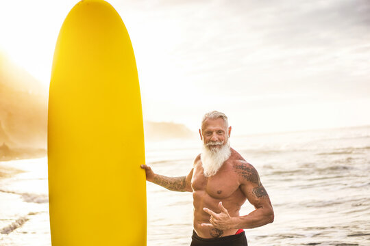 Tattooed senior surfer holding surf board on the beach at sunset - Happy old guy having fun doing extreme sport - Joyful elderly concept - Focus on his face - Powered by Adobe