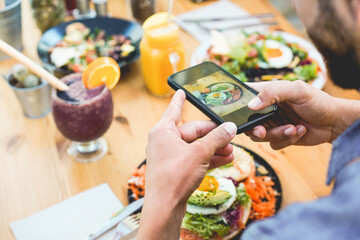 Naklejka na ściany i meble Influencer man eating brunch while making video and photos of dish with mobile phone in trendy bar restaurant - Healthy lifestyle, technology and food trends concept - Focus on man hand