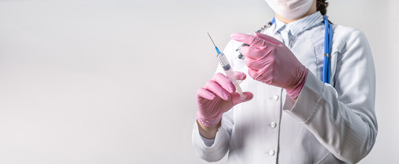 Cropped view of young female doctor in a white coat, in a medical mask and pink sterile gloves hold syringe. Focus at syringe. Vaccination concept.