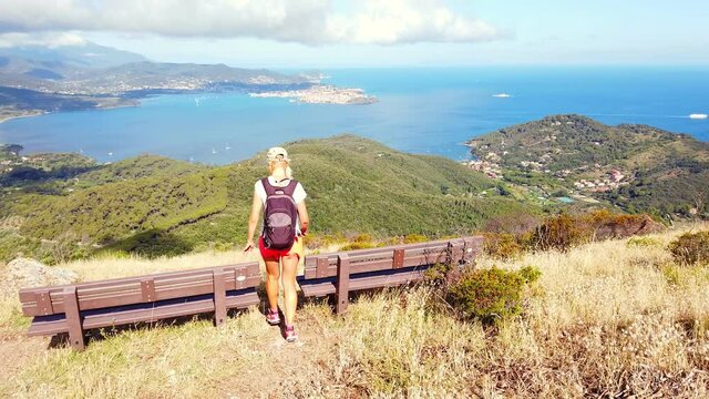 Tourist woman backpacker on benches for relax during trekking to Volterraio Castle and looking panoramic views of Portoferraio Gulf, Elba Island. Tourism in Tuscany, Italy travel destination, Europe.