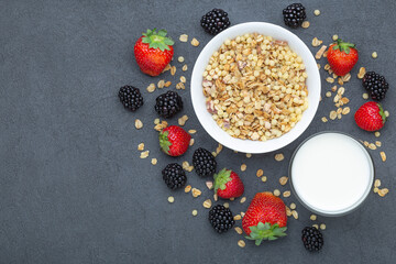 Healthy breakfast ingredients. Homemade granola in white plate, milk or yogurt in glass, blackberries and strawberries on stone background, view from above, copy space.