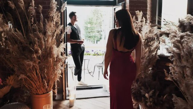 Back view at sexy girlfriend going to romantic evening meeting to waiting boyfriend standing on luxury restaurant balcony indoor. Passionate romance, love feelings of young elegant affectionate couple