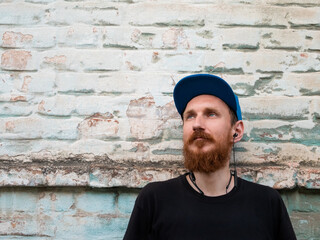 Portrait of young red bearded man in black t-shirt and snapback in rock style on urban background listening to music with headphones. Hipster guy in wireless earphones on old blue brick wall backdrop.
