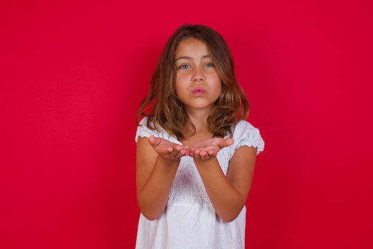 Little caucasian girl with blue eyes wearing white dress standing over isolated red background sending blow kiss with pout lips and holding palms to send air kiss.