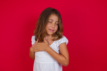 Little caucasian girl with blue eyes wearing white dress standing over isolated red background smiling with hands on chest with closed eyes and grateful gesture on face. Health concept.