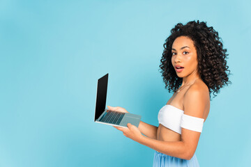 surprised and curly african american woman holding laptop isolated on blue