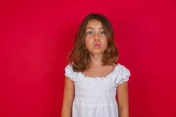 Shot of pleasant looking Little caucasian girl with blue eyes wearing white dress standing , pouts lips, looks with green eyes at camera,  has fun with girlfriend, Human facial expressions