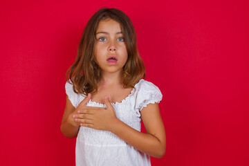 Scared Little caucasian girl with blue eyes wearing white dress looks with frightened expression, keeps hands on chest, being puzzled to notice something strange,  People, hush reaction and emotions.