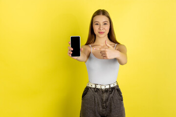 Naklejka premium Pointing on phone's screen. Caucasian young girl's portrait isolated on yellow studio background. Beautiful female blonde model. Concept of human emotions, facial expression, sales, ad, youth culture
