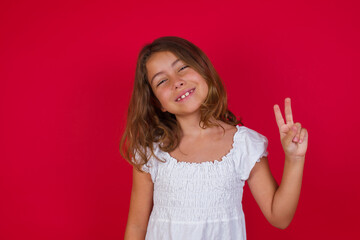 Little caucasian girl with blue eyes wearing white dress standing over isolated red background smiling with happy face winking at the camera doing victory sign. Number two.