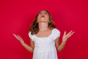 Little caucasian girl with blue eyes wearing white dress standing over isolated red background crazy and mad shouting and yelling with aggressive expression and arms raised. Frustration concept.