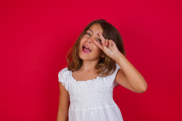 Little caucasian girl with blue eyes wearing white dress standing over isolated red background Doing peace symbol with fingers over face, smiling cheerful showing victory