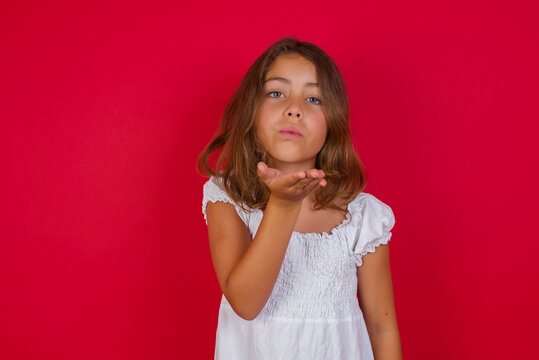 Little caucasian girl with blue eyes wearing white dress standing over isolated red background looking at the camera blowing a kiss with hand on air being lovely and sexy. Love expression.