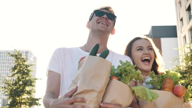 Happy carefree couple laughs walking along street to home from market, carrying bags with fresh healthy food. Fun grocery shopping together, casual life of young excited family enjoying joke outdoors