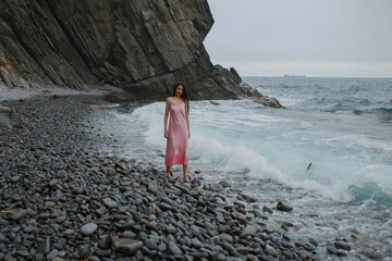 A dark-haired girl with long hair in a pink dress stands on a stone beach near the blue sea on a background of high steep cliffs