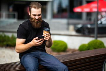 Handsome young brutal bearded man sitting on bench and checking his smart phone outdoors