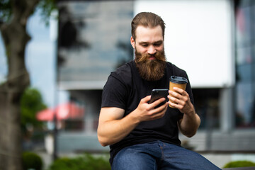 Handsome young brutal bearded man sitting on bench and checking his smart phone outdoors