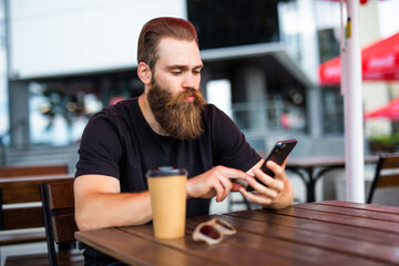 Portrait of serious young man with the beard texting message into his workgroup using the smartphone while waiting for a food order in street restaurant