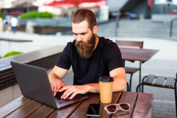 Young man wearing eyeglasses holding cup of coffee and typing on laptop while working in city cafe...