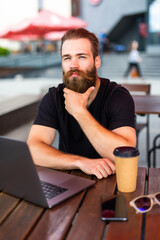 Young man wearing eyeglasses holding cup of coffee and typing on laptop while working in city cafe...