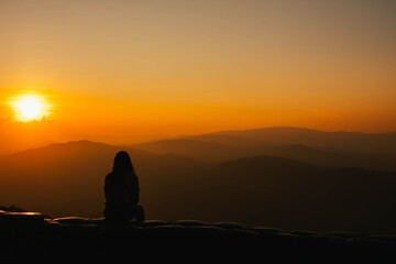 Silhouette of a girl sitting on a cliff side looking at the sunset.  Beautiful sunset background.