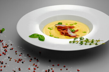 Corn cream soup with smoked meat in a white plate decorated with basil, on a gray background. The first course on the restaurant menu.