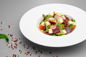 Vegetable salad with radish, soft cheese, beetroot and greens in a white plate on a gray background. Snacks on the restaurant.