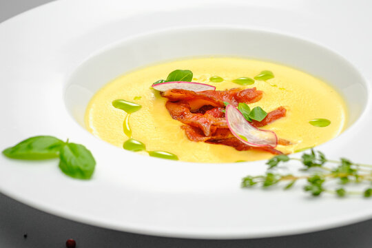 Corn cream soup with smoked meat in a white plate decorated with basil, on a gray background. The first course on the restaurant menu.