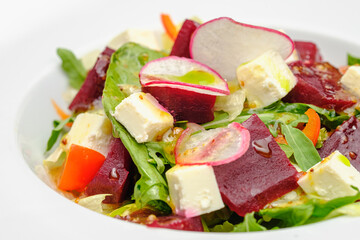 Vegetable salad with radish, soft cheese, beetroot and greens in a white plate on a gray background. Snacks on the restaurant.