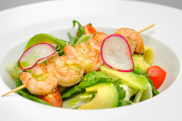 Grilled shrimp on a skewer with vegetables on a white plate on a gray background.