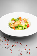 Grilled shrimp on a skewer with vegetables on a white plate on a gray background.