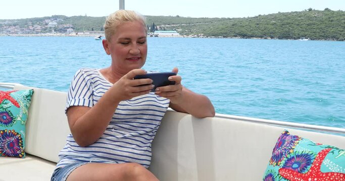 Portrait of a middle aged beautiful woman is sitting on the deck of a yacht with a smartphone at summer vacation