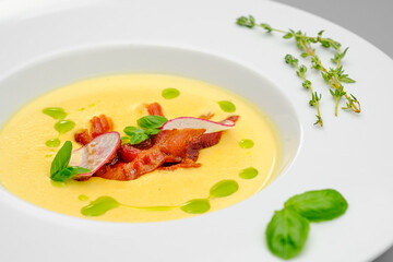 Corn cream soup with smoked meat in a white plate decorated with basil, on a gray background. The first course on the restaurant menu.