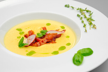Corn cream soup with smoked meat in a white plate decorated with basil, on a gray background. The first course on the restaurant menu.