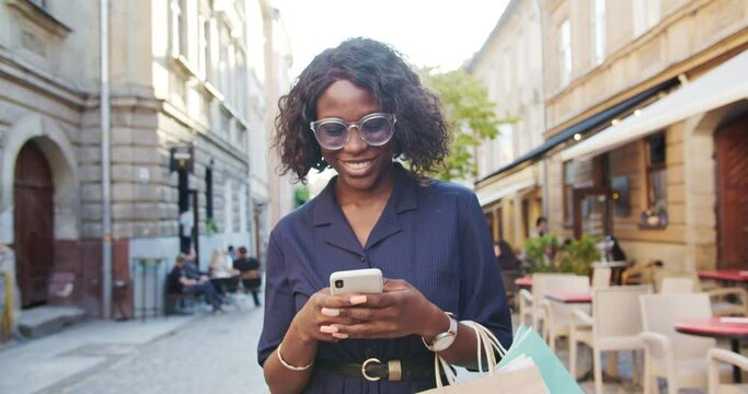 Close up portrait of joyful pretty African American young woman texting on smartphone while walking in town. Beautiful girl with shopping bags smiling while typing on cellphone outdoor. City concept