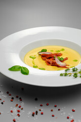 Corn cream soup with smoked meat in a white plate decorated with basil, on a gray background. The first course on the restaurant menu.