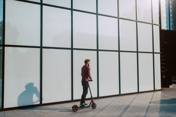 A handsome young bearded businessman rides  e scooter in front of the white wall of the building