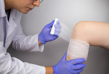 The doctor bandages the knee of a girl who is injured. Stopping bleeding by applying a pressure bandage to the injury. First aid for injuries. The technique of applying a cruciform bandage.