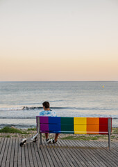 Boy sitting with his scooter on a bench painted in the colors of gay pride overlooking the sea.