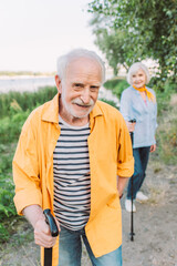 Selective focus of smiling senior man with walking stick looking at camera near wife in park