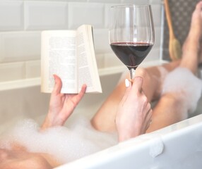 Woman holding a glass of red wine and reading book in bath with foam. Concepts of unrecognizable...