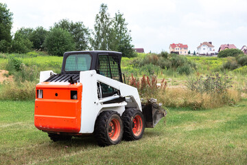 A skid steer loader clears the site for construction. Land work by the territory improvement. Machine for work in confined areas. Small tractor with a bucket for moving soil and bulk materials.