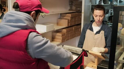 Handheld shot of cheerful female coffeeshop employee in apron carrying boxes and handing them to delivery man with insulated backpack